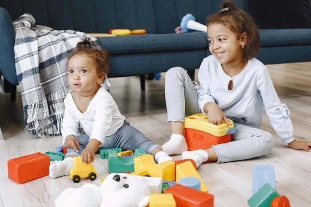 two pretty kids sit on floor and play with toys near sofa. afro-american sisters playing at home.